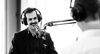 A man is sitting in front of a microphone with headphones on, speaking for a podcast.